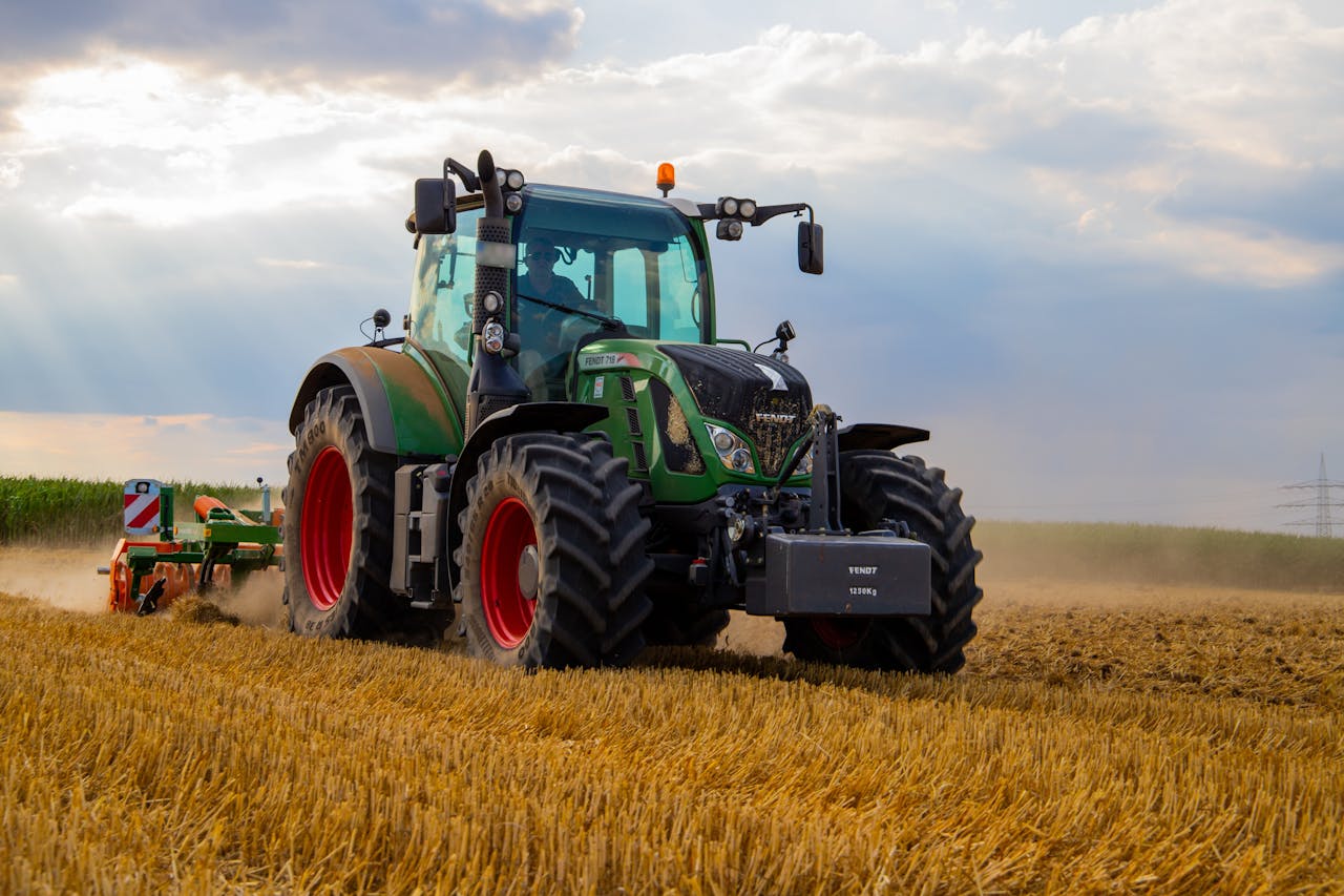 who-we-are A powerful green tractor plowing a dusty wheat field under a cloudy summer sky in rural Germany.